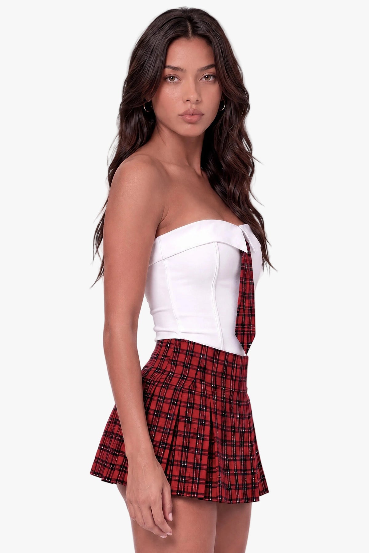 A woman modeling a strapless white top with a red and black plaid bow detail, paired with a matching red and black plaid pleated mini skirt, against a light gray studio background.