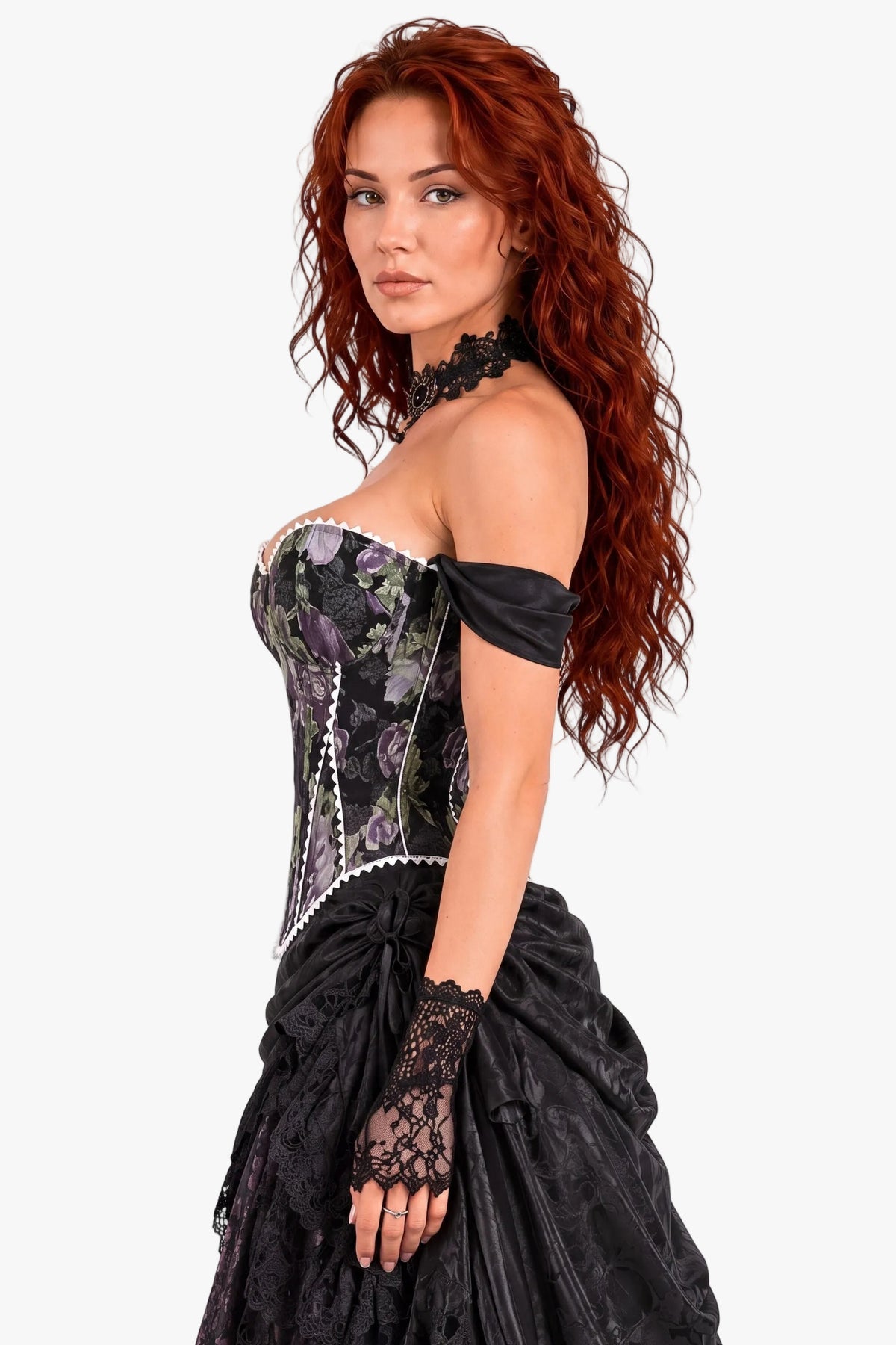 A woman with long, curly red hair models a Victorian-style off-the-shoulder dress featuring a floral-patterned corset bodice, black lace gloves, and a voluminous black skirt against a plain white studio background.
