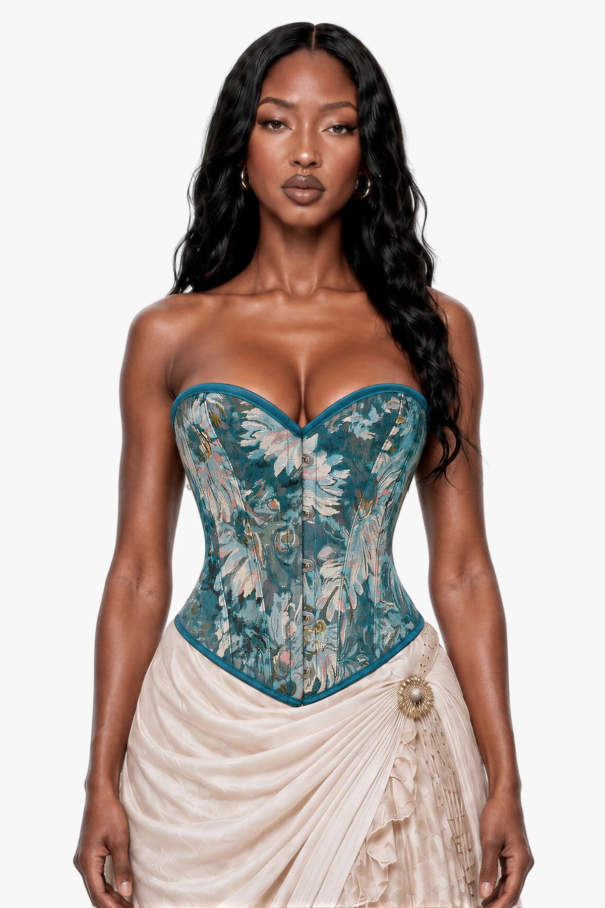 A woman modeling a strapless corset top with a teal blue base and a white floral pattern, paired with a cream draped skirt, against a light gray studio background.