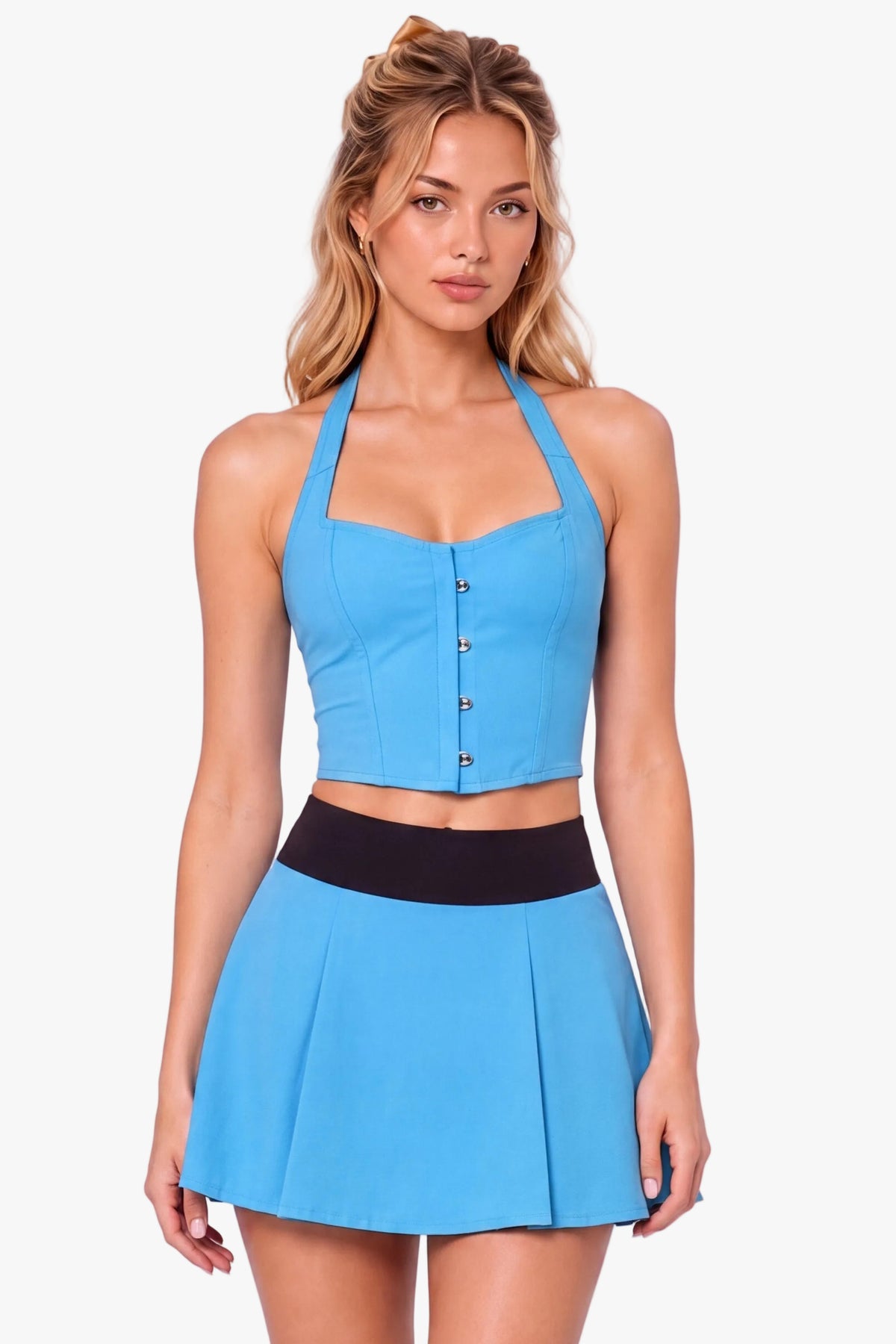 A woman modeling a bright blue halter crop corset top and matching pleated mini skirt set with a black waistband, styled with white socks and black Mary Jane shoes against a light lavender studio background.