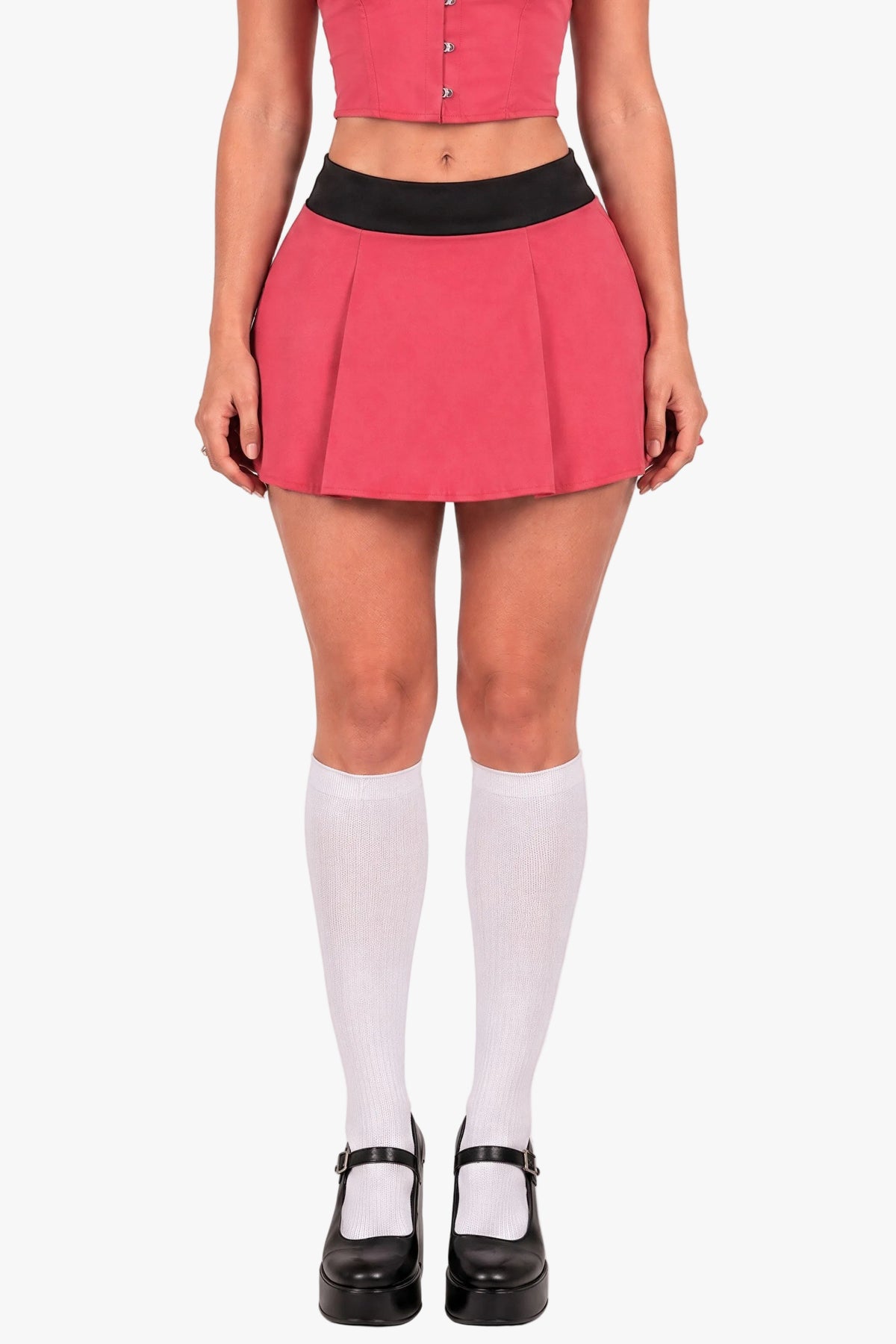 A model is shown from the waist down wearing a bright pink pleated mini skirt with a black waistband, paired with white knee-high socks and black Mary Jane shoes, against a plain light gray studio background.