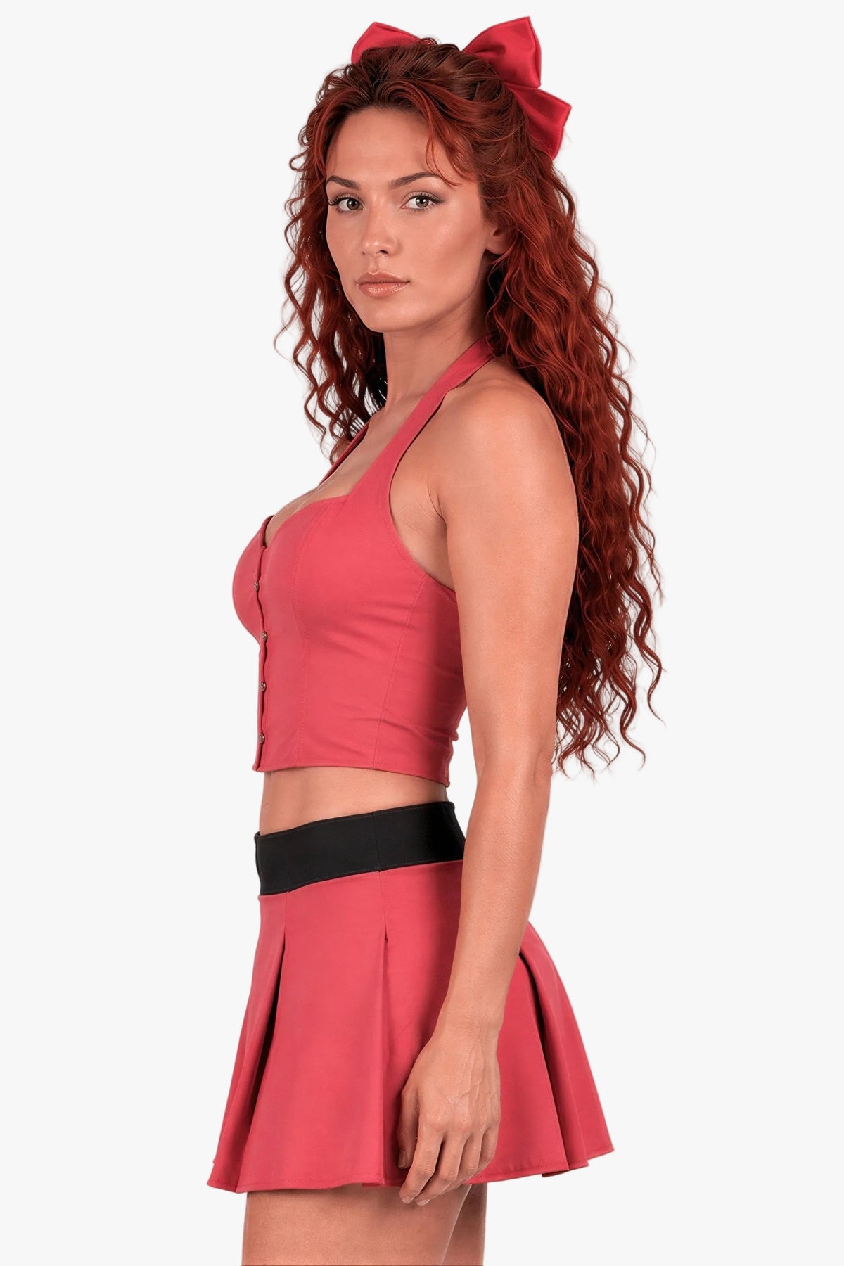 A woman modeling a powerpuff girl blossom costume - a two-piece costume set consisting of a coral pink halter crop top and matching pleated mini skirt with a black waistband, plus a matching red bow headband. She is photographed against a plain light gray studio background.