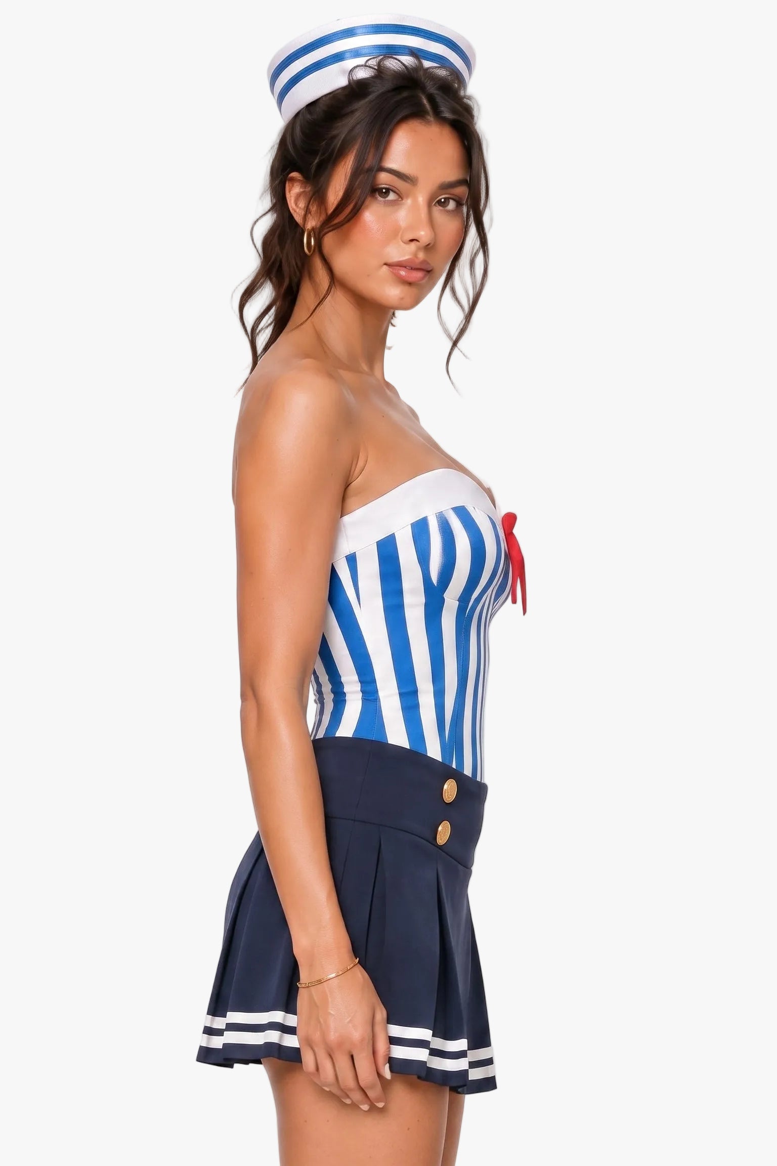 A woman modeling a sailor-themed costume set featuring a blue and white striped strapless top with a red bow, a navy blue pleated mini skirt with white stripes, and a matching navy blue sailor hat, against a plain light gray studio background.