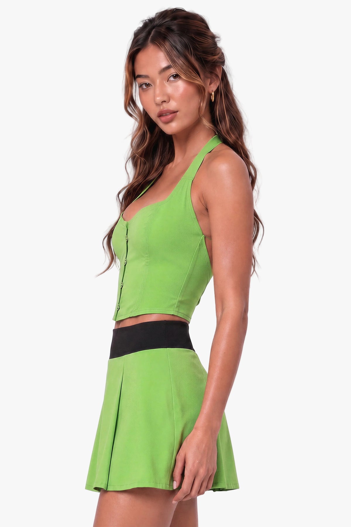 A woman modeling a bright lime green two-piece costume set consisting of a halter crop corset top and a matching pleated mini skirt with a wide black waistband, set against a solid light lavender studio background.