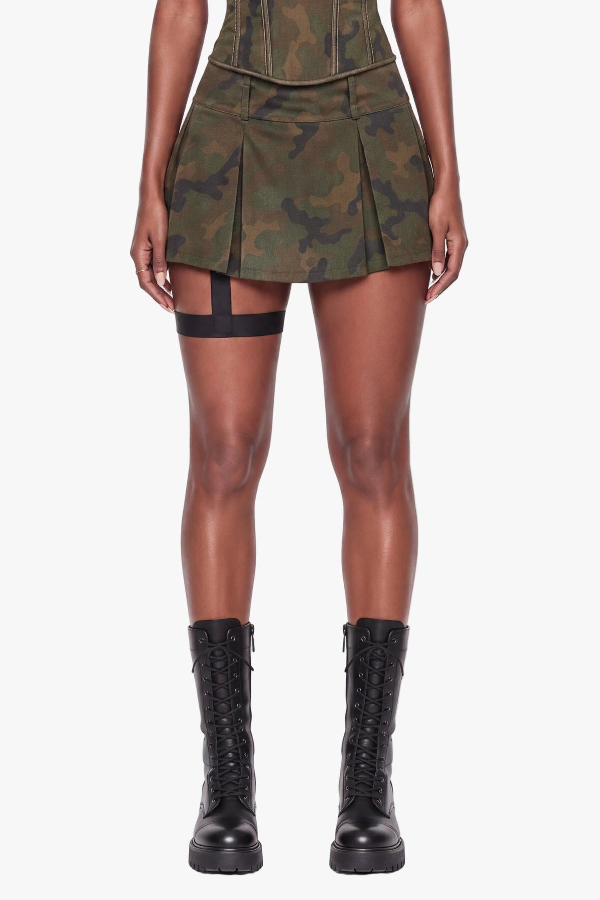 A model stands against a plain white studio background wearing a strapless mini dress with a green camouflage pattern, structured corset-style bodice, and pleated skirt, paired with black lace-up boots.