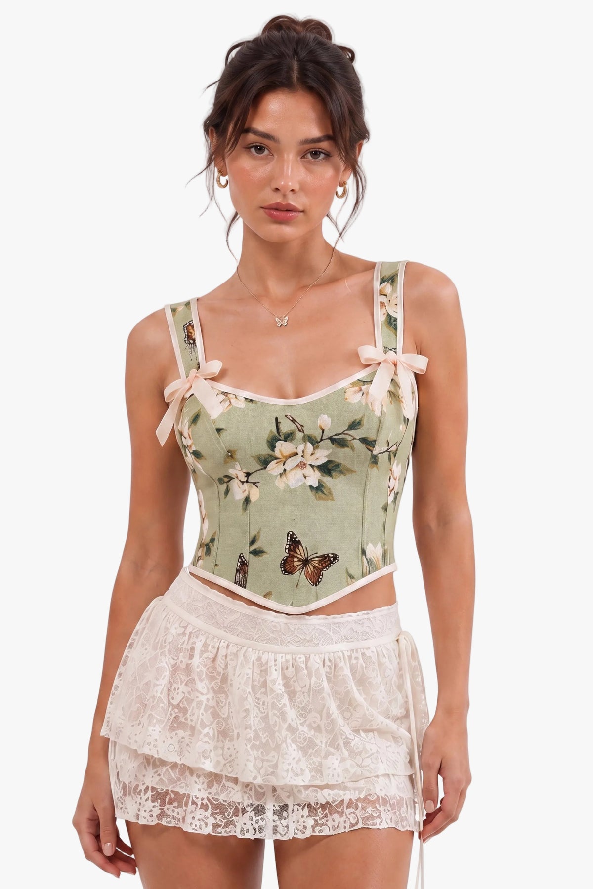 A woman modeling a light sage green floral corset top with butterfly prints and cream lace ruffled shorts against a plain white background.
