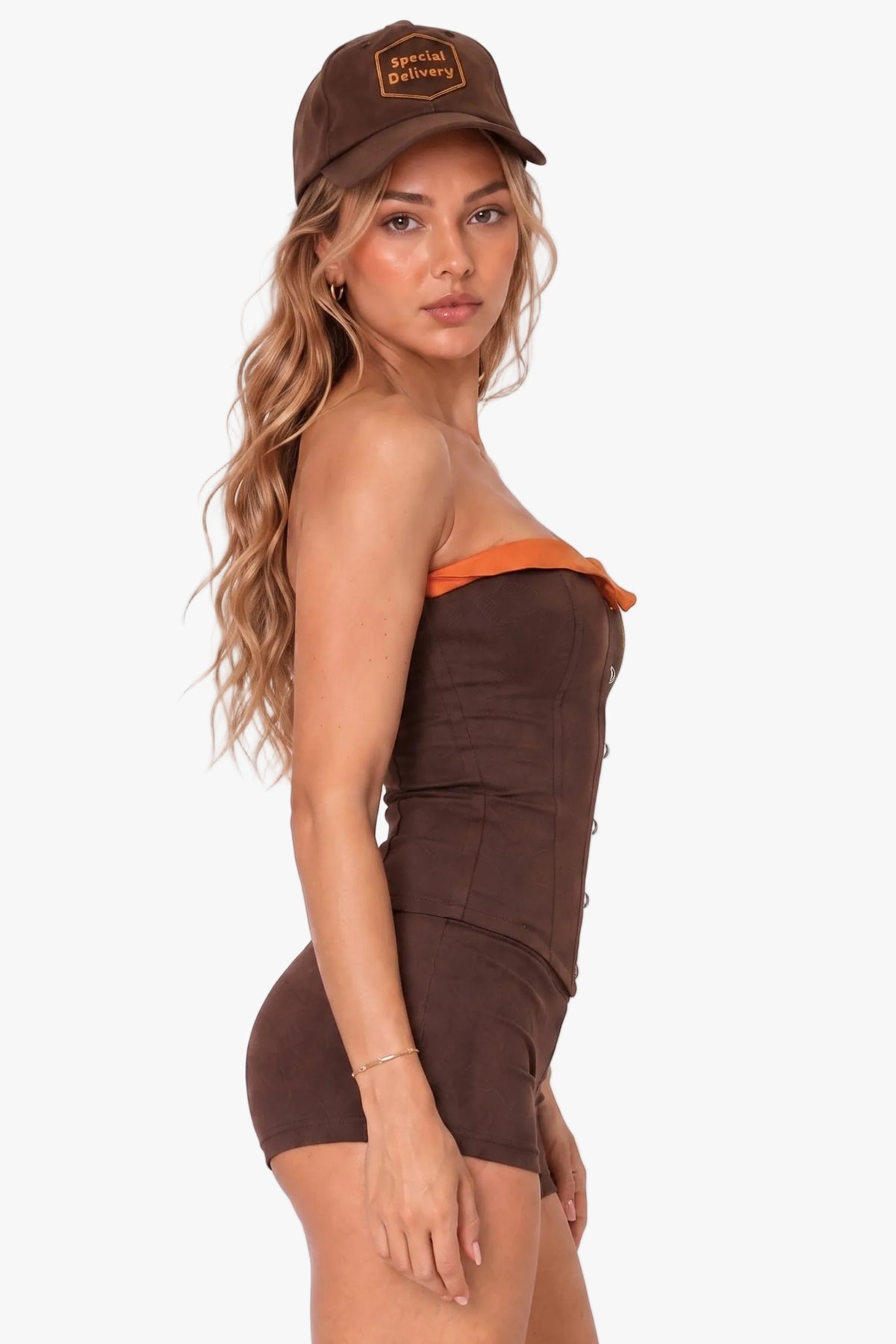 A woman modeling a brown strapless corset with orange trim and a matching brown baseball cap, photographed against a plain light background.