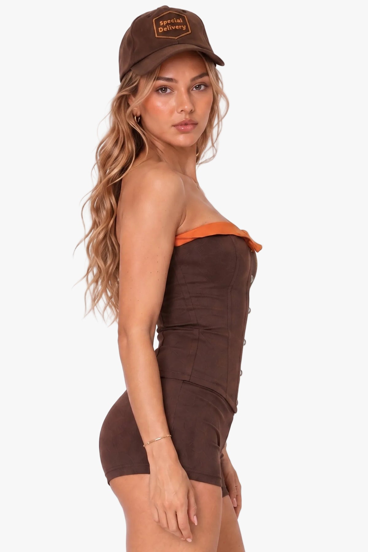 A woman modeling a brown strapless corset with orange trim and a matching brown baseball cap, photographed against a plain light background.