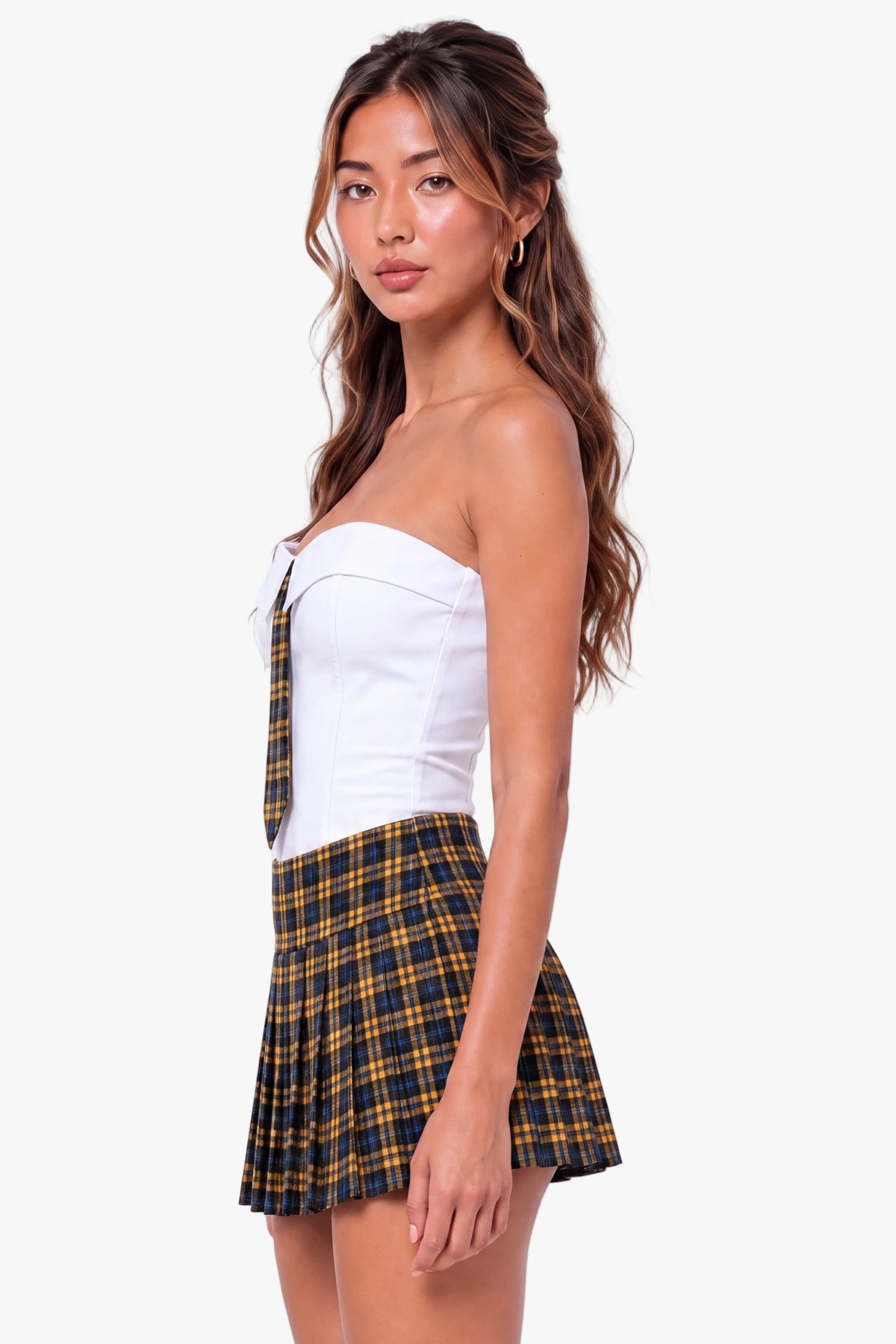 A woman modeling a white strapless tube top and a black and yellow plaid pleated mini skirt against a plain white studio background.
