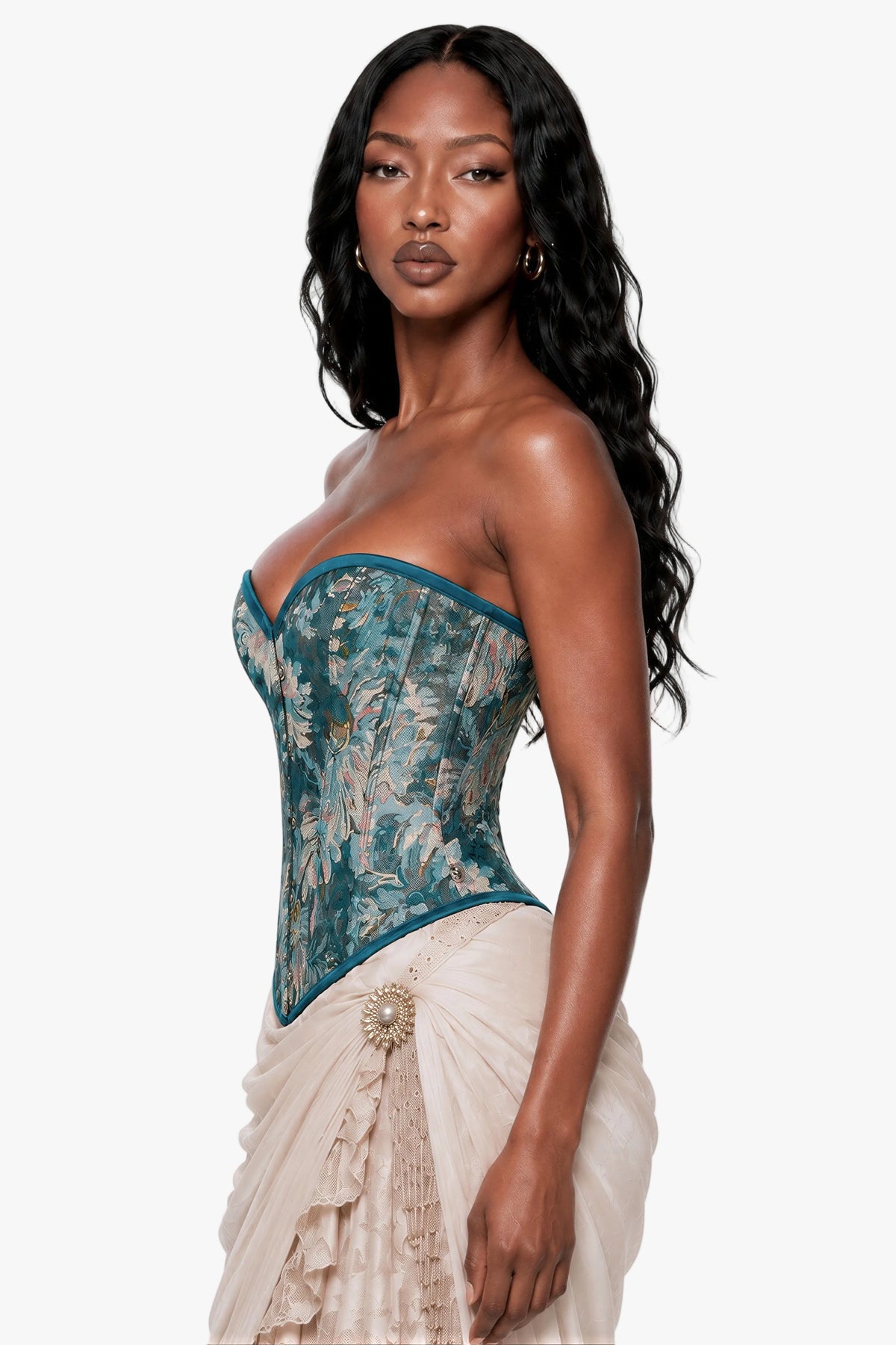 A woman modeling a strapless corset top with a teal and beige floral lace overlay, paired with a light beige ruffled skirt, against a plain light gray studio background.