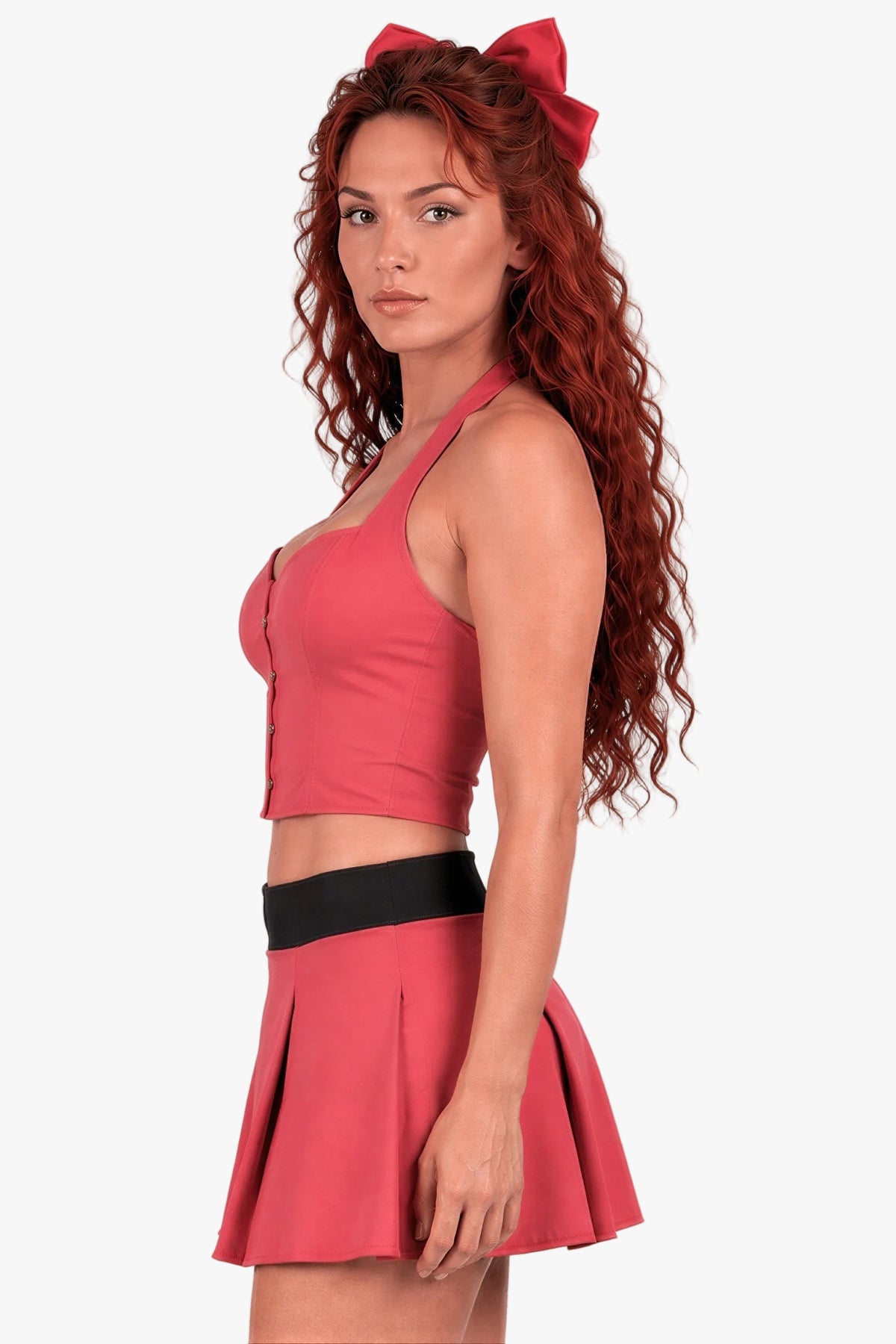 A woman with long curly red hair wearing a matching red two-piece outfit with a halter crop corset top, pleated mini skirt, and a large red bow headband, posed against a light gray studio background.