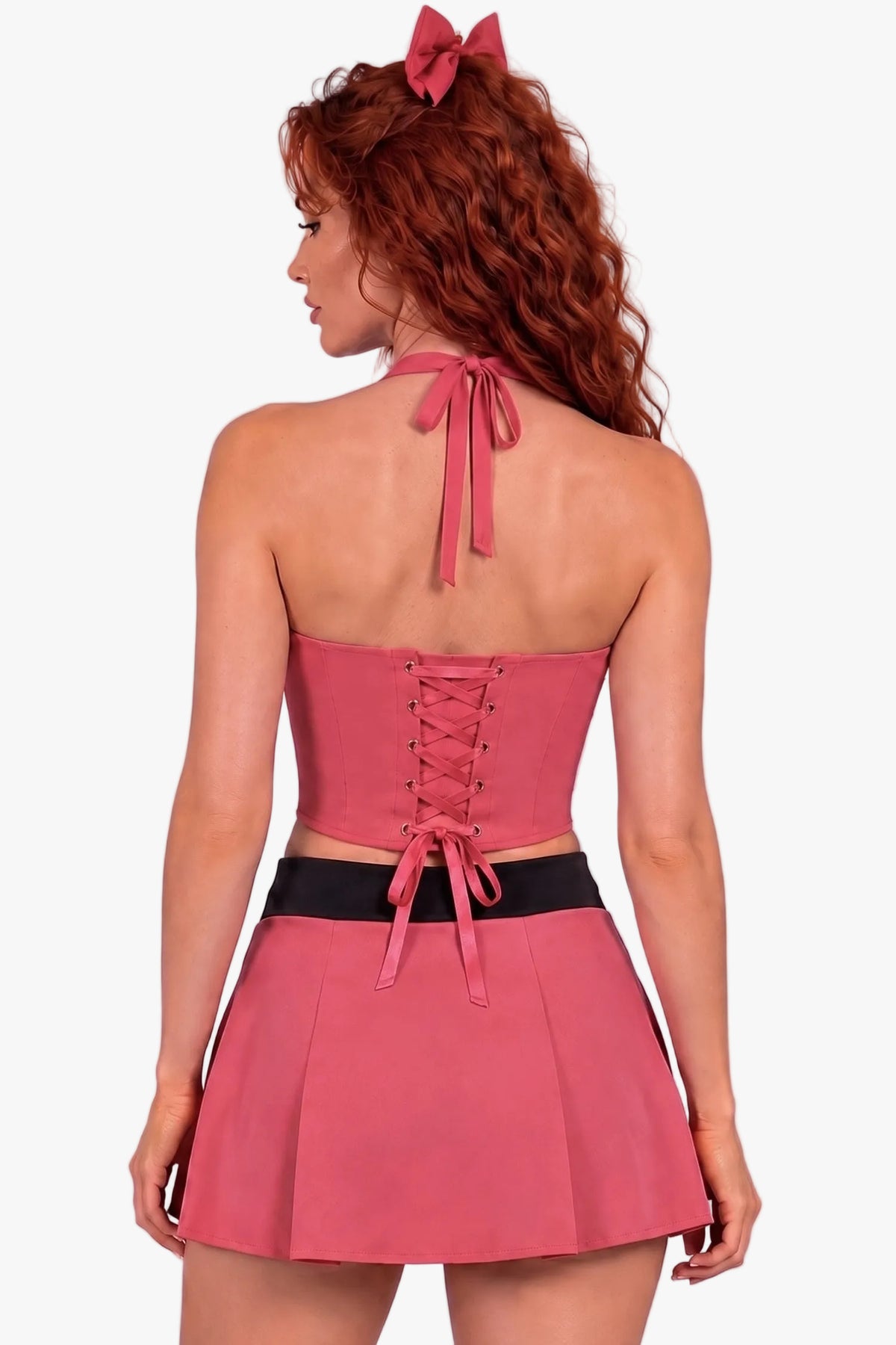 A woman with red hair wearing a pink halter corset and a skirt with a black waistband, shown from the back against a solid lavender background.