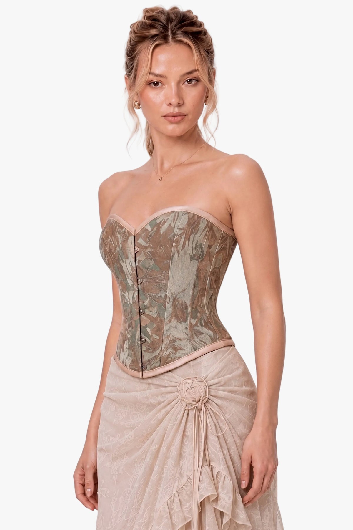 A woman modeling a strapless corset-style top with a muted floral camouflage pattern in beige, taupe, and brown tones, paired with a matching draped skirt against a plain light gray studio background.