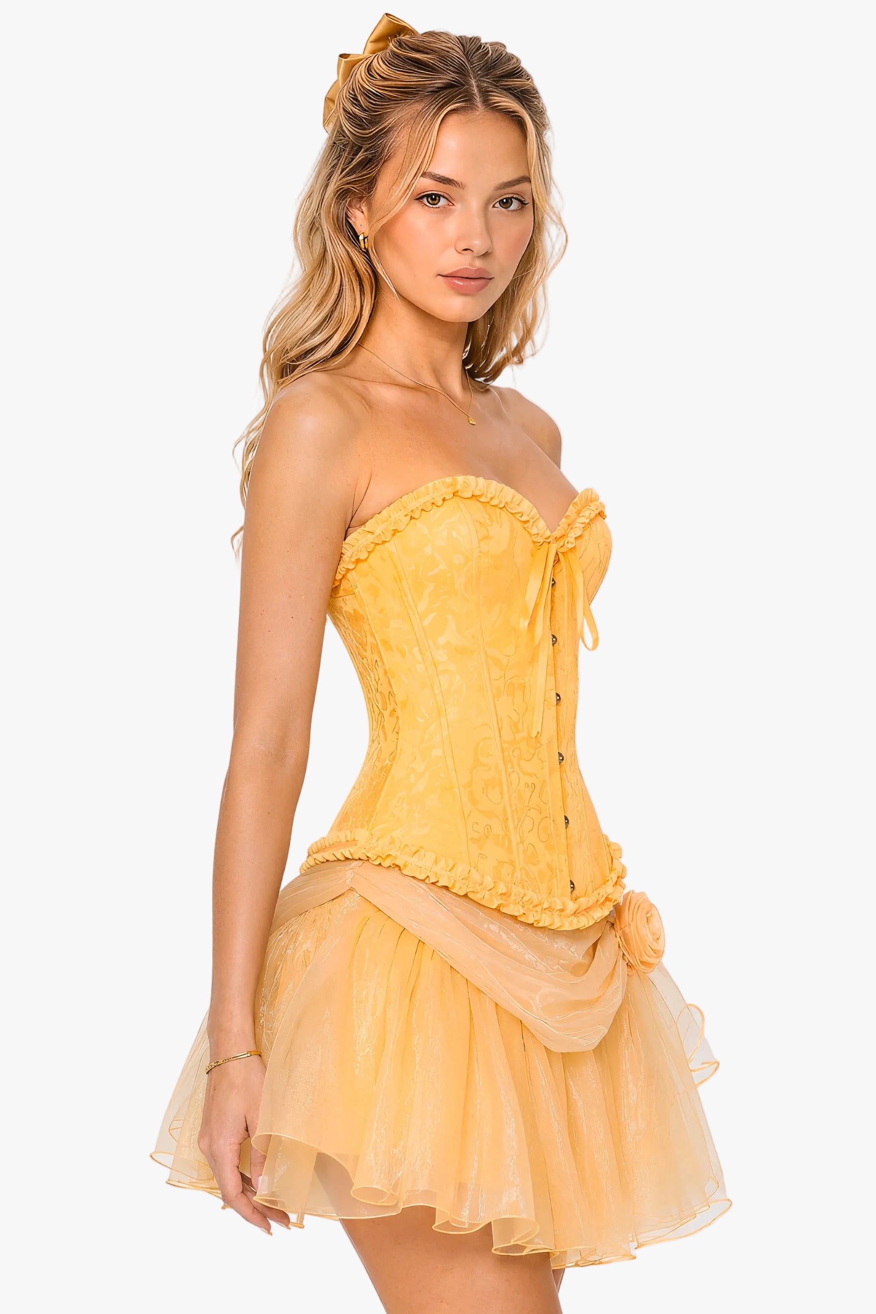 A woman modeling a yellow strapless corset mini dress with a lace-embroidered bodice, ruffled trim, front tie, and a layered tulle skirt, photographed against a plain light gray studio background.