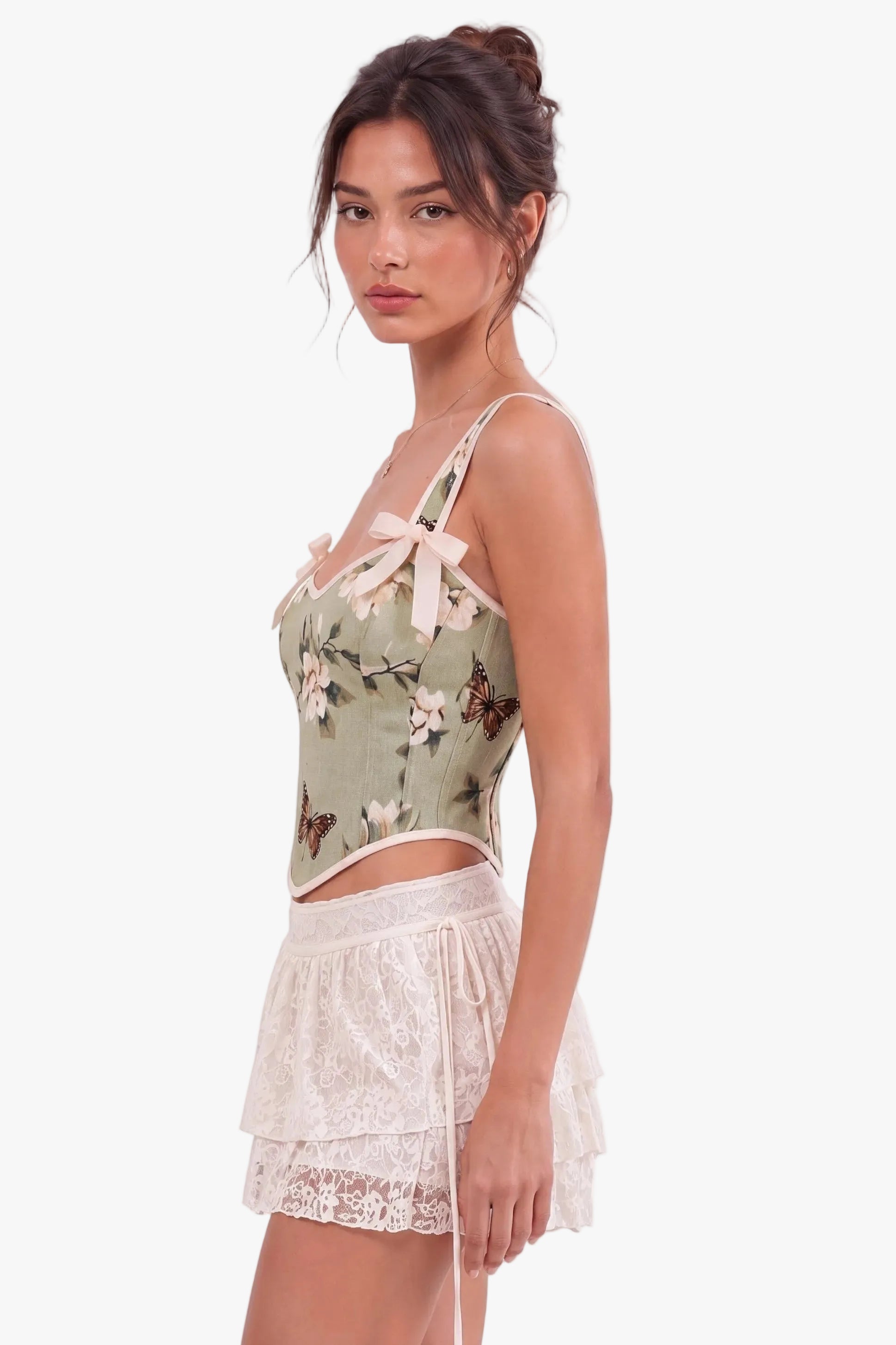 A woman modeling a light sage green camisole top with a butterfly and floral print, paired with a white lace tiered mini skirt, against a plain white background.