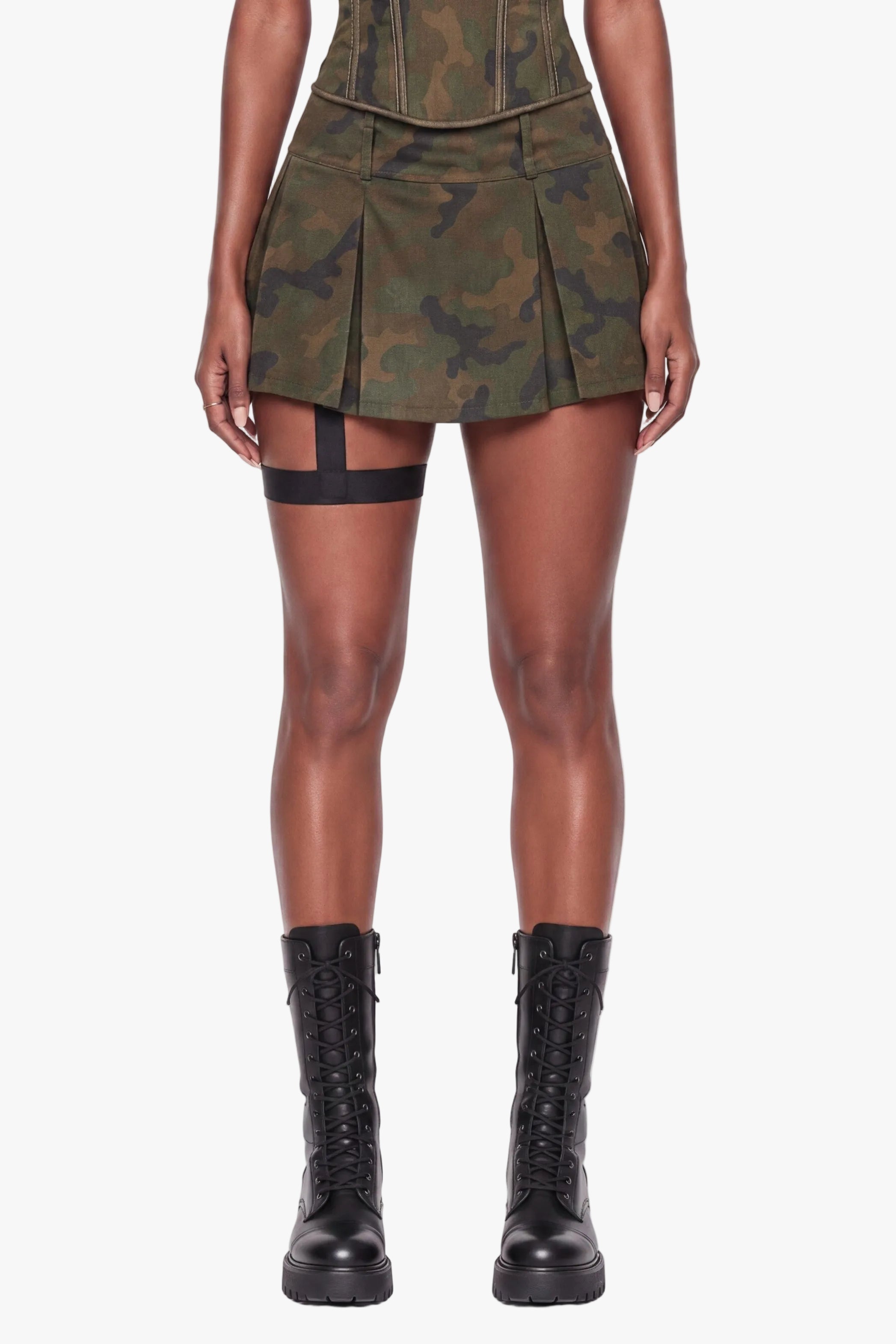 A model stands against a plain white studio background wearing a strapless mini dress with a green camouflage pattern, structured corset-style bodice, and pleated skirt, paired with black lace-up boots.