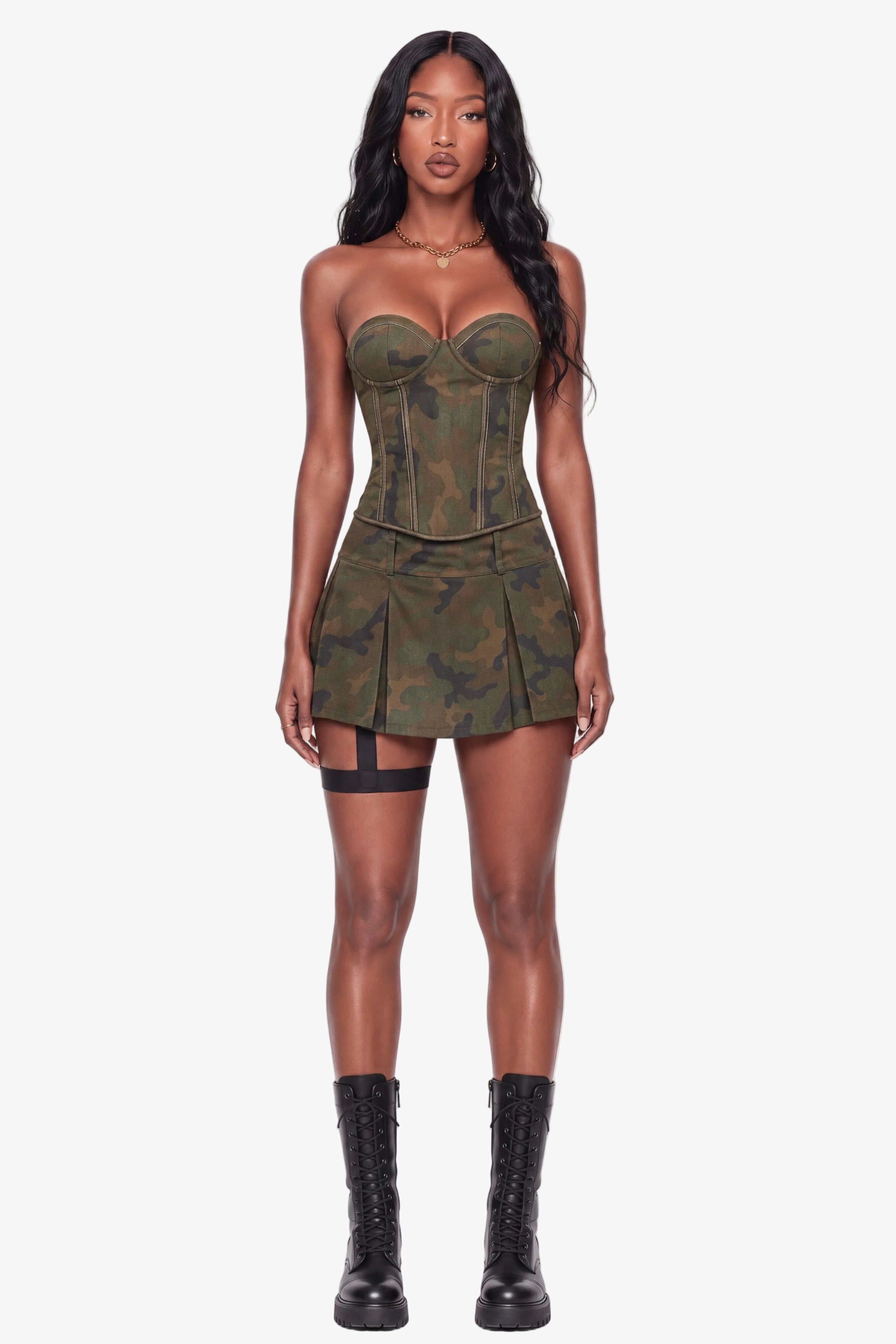 A model stands against a plain white studio background wearing a strapless mini dress with a green camouflage pattern, structured corset-style bodice, and pleated skirt, paired with black lace-up boots.
