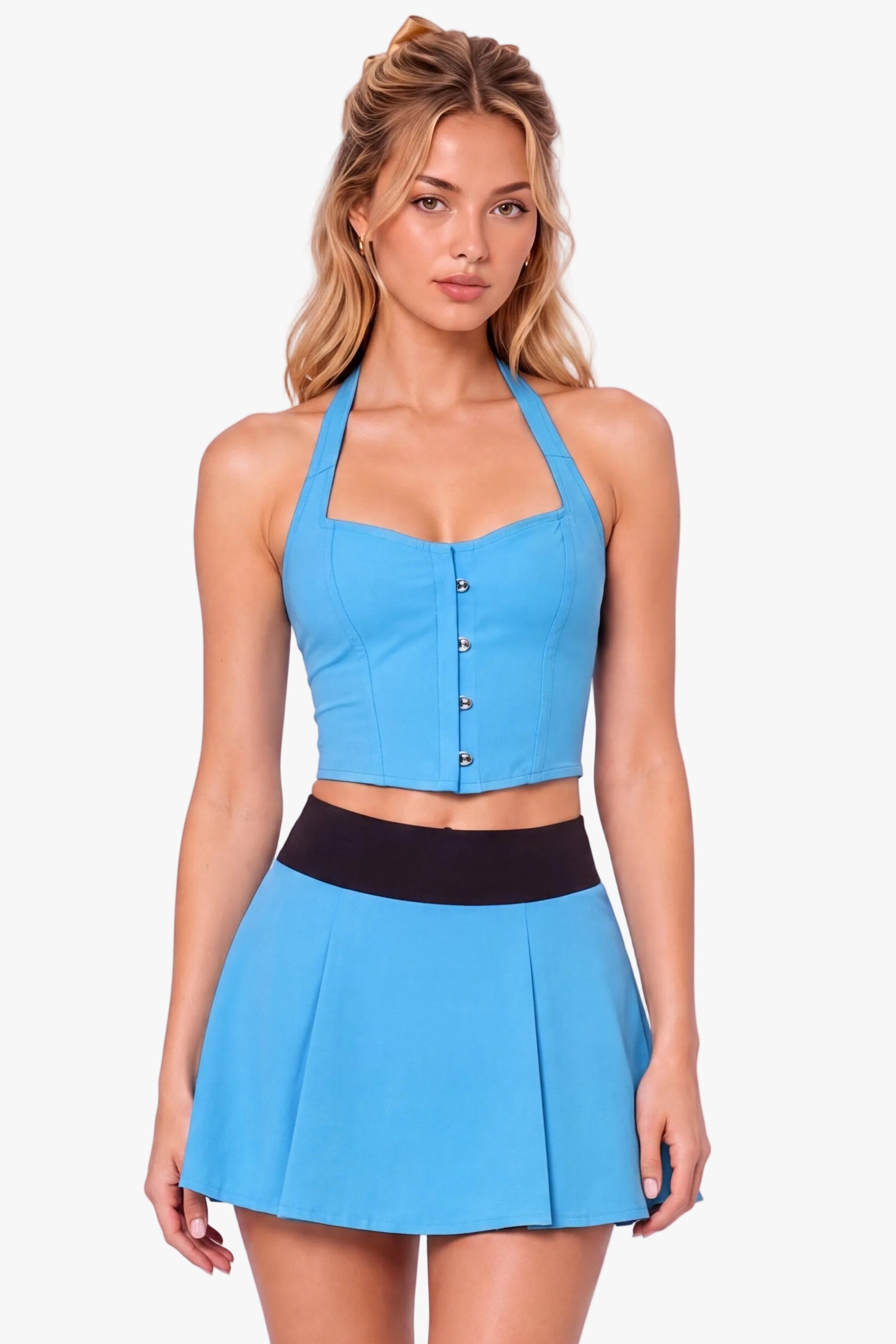 A woman modeling a bright blue halter crop top and matching pleated mini skirt set with a black waistband, styled with white socks and black Mary Jane shoes against a light lavender studio background.