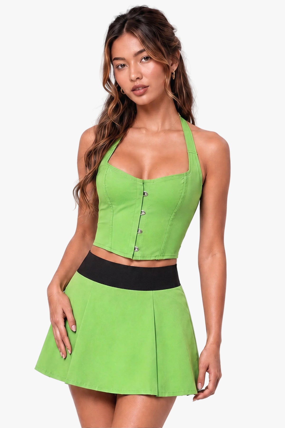 A woman modeling a bright lime green two-piece costume set consisting of a halter crop corset top and a matching pleated mini skirt with a wide black waistband, set against a solid light lavender studio background.