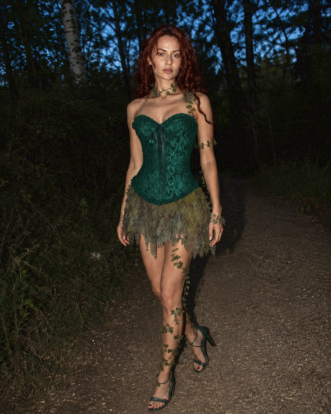 A woman with red hair stands on a forest path at dusk wearing a dark green lace corset top, dressed up as poison ivy, with a matching feathered mini skirt, and green lace-up heels, with floral vine graphics on her outfit.